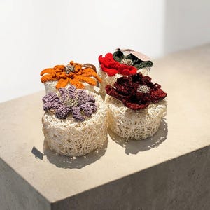 May include: Four handmade decorative objects, each featuring a unique crocheted flower design atop a woven base. The flowers are in shades of orange, red, purple, and burgundy. The objects are displayed on a neutral-toned surface.