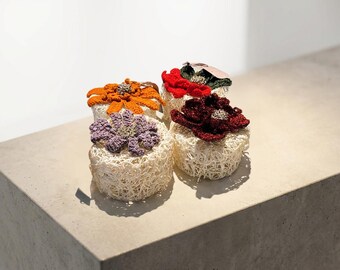 Puede incluir: Cuatro objetos decorativos hechos a mano, cada uno con un diseño floral único tejido a ganchillo sobre una base tejida. Las flores son en tonos naranja, rojo, morado y burdeos. Se exhiben sobre una superficie neutra.