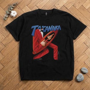 Könnte beinhalten: Ein schwarzes Kurzarm-T-Shirt mit einem Grafikdruck einer Figur im roten Anzug, die eine rote Gitarre spielt. Über der Figur ist der Text TAKANAKA in Blau gedruckt. Das T-Shirt hat einen Rundhalsausschnitt und eine klassische Passform.