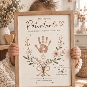 May include: A rectangular wooden frame containing a white print with the German text "FÜR MEINE Patentante DANKE, DASS DU IMMER FÜR MICH DA BIST." The print features a central handprint surrounded by floral illustrations and a bow.