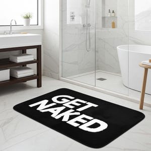 Può includere: Un tappetino da bagno rettangolare con uno sfondo nero a tinta unita e la scritta GET NAKED stampata in grandi lettere maiuscole bianche e in grassetto al centro. Il tappetino ha angoli arrotondati e una consistenza morbida e felpata.