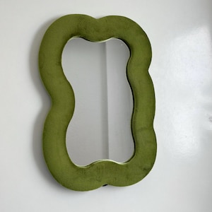 May include: A wall mirror featuring a wavy, organic-shaped frame covered in a soft, olive green velvet fabric. The central reflective glass surface follows the irregular, undulating contours of the plush, textured border.