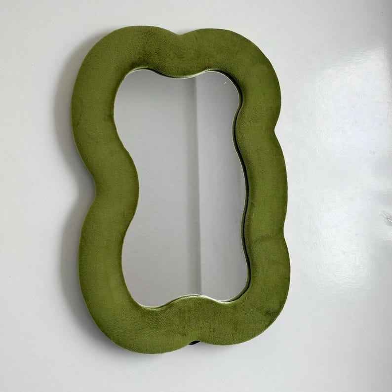 May include: A wall mirror featuring a wavy, organic-shaped frame covered in a soft, olive green velvet fabric. The central reflective glass surface follows the irregular, undulating contours of the plush, textured border.
