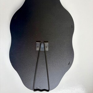 May include: The back of a black, scalloped-edge frame featuring a central metal easel stand and a top-mounted metal hanging hook. The dark backing is secured with small metal staples along the perimeter, providing a sturdy structure for display.