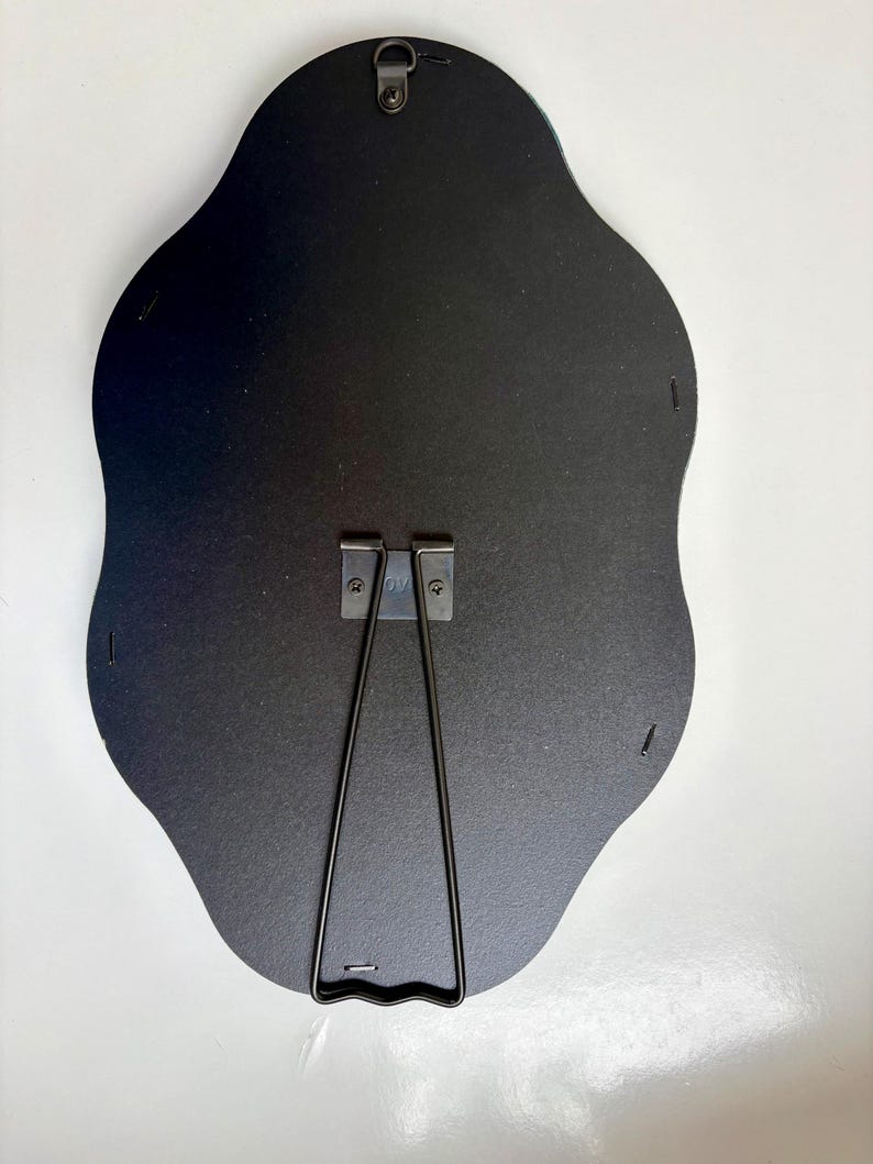 May include: The back of a black, scalloped-edge frame featuring a central metal easel stand and a top-mounted metal hanging hook. The dark backing is secured with small metal staples along the perimeter, providing a sturdy structure for display.