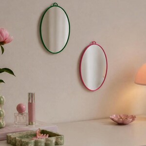 Pode incluir: Dois espelhos de parede ovais com molduras finas, um em verde escuro e outro em rosa brilhante, cada um com um pequeno laço circular para pendurar no topo. Os espelhos estão montados numa parede lisa sobre uma superfície com objetos.