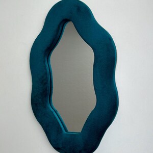 May include: A wall mirror featuring an irregular, wavy-shaped frame covered in a deep teal velvet fabric. The reflective glass surface follows the organic, undulating contours of the soft, plush frame, creating a distinct, non-symmetrical silhouette.