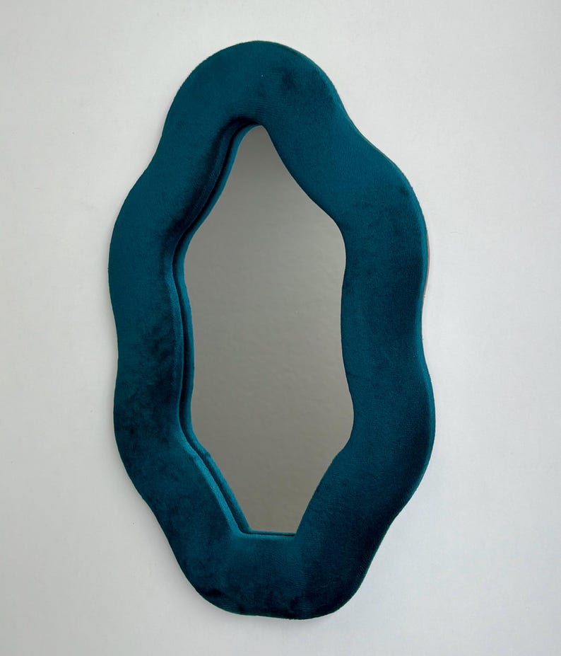 May include: A wall mirror featuring an irregular, wavy-shaped frame covered in a deep teal velvet fabric. The reflective glass surface follows the organic, undulating contours of the soft, plush frame, creating a distinct, non-symmetrical silhouette.
