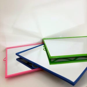 Op de afbeelding: Drie kleine, rechthoekige spiegels met gekleurde frames. Eén heeft een roze frame, één heeft een blauw frame en de derde heeft een groen frame. Elke spiegel heeft aan één kant een klein handvat.