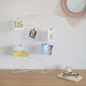 Può includere: Un espositore fotografico in metallo bianco con un design geometrico, che contiene diverse piccole foto fissate con clip. L'espositore è su una superficie di legno, con uno specchio, un libro e occhiali sullo sfondo.