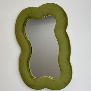 May include: A wall mirror featuring an irregular, wavy-shaped frame covered in a soft, olive green velvet fabric. The central reflective glass surface follows the same organic, undulating contour as the plush, textured outer border.