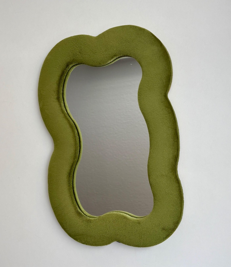 May include: A wall mirror featuring an irregular, wavy-shaped frame covered in a soft, olive green velvet fabric. The central reflective glass surface follows the same organic, undulating contour as the plush, textured outer border.