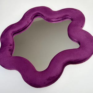 May include: A wall mirror featuring a thick, wavy, organic-shaped frame covered in a deep purple velvet fabric. The reflective glass surface is centered within the plush, soft-textured border, creating a distinct contrast between the smooth mirror and the fabric.
