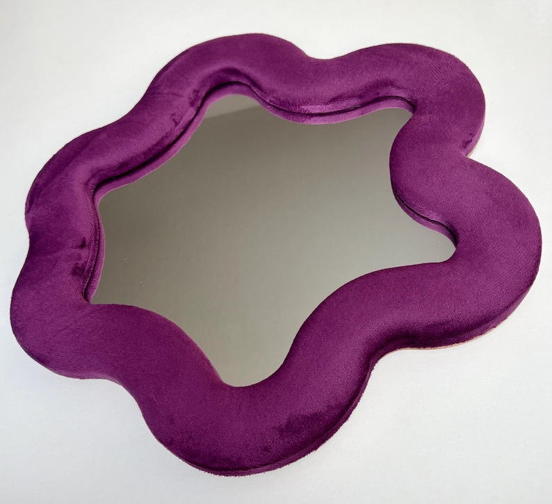 May include: A wall mirror featuring a thick, wavy, organic-shaped frame covered in a deep purple velvet fabric. The reflective glass surface is centered within the plush, soft-textured border, creating a distinct contrast between the smooth mirror and the fabric.