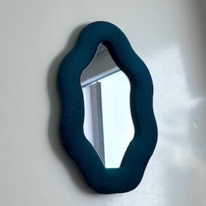 May include: A wall-mounted mirror featuring a thick, wavy frame upholstered in a deep teal velvet fabric. The central reflective glass surface follows the organic, undulating shape of the frame, creating a soft, rounded silhouette for the piece.
