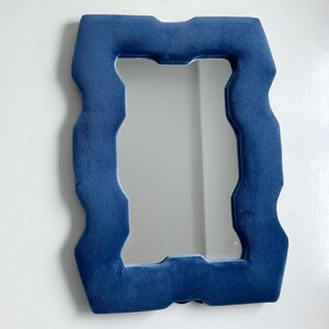 May include: A rectangular wall mirror featuring a thick, plush, deep blue velvet frame with a distinct wavy, undulating edge design. The reflective glass surface is centered within the soft, textured blue border, creating a bold contrast.