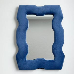 May include: A rectangular mirror featuring a thick, plush, deep blue velvet frame with a distinct wavy, undulating edge design. The reflective glass surface is centered within the soft, textured border, creating a bold contrast between the smooth mirror and the fabric frame.