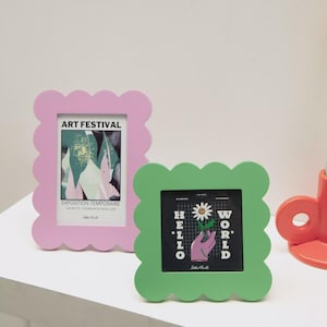 Peut inclure: Deux cadres photo décoratifs aux bords festonnés. L'un est rose clair et affiche une affiche de festival d'art. L'autre est vert et présente un graphisme avec les mots "Hello World" et une main tenant une fleur.
