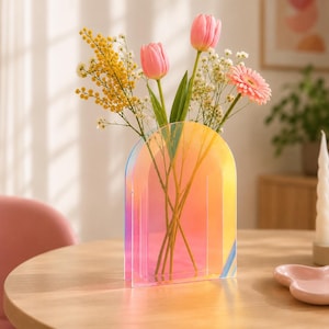 Può includere: Un vaso ad arco in acrilico iridescente con una sfumatura di tonalità rosa, arancione e gialle. Il vaso contiene una composizione floreale con due tulipani rosa, un rametto di mimosa gialla, nebbiolina bianca e una singola gerbera rosa.