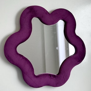 May include: A wall-mounted mirror featuring a thick, plush, deep purple velvet frame with a distinct wavy, organic flower-like shape. The central reflective glass surface follows the same undulating contour as the soft, textured fabric border.