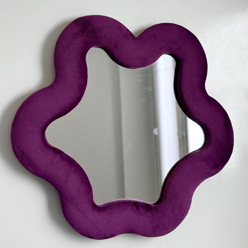 May include: A wall-mounted mirror featuring a thick, plush, deep purple velvet frame with a distinct wavy, organic flower-like shape. The central reflective glass surface follows the same undulating contour as the soft, textured fabric border.