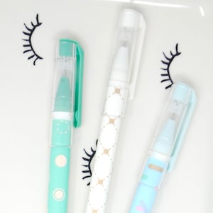 May include: Three colorful gel pens with clear caps. One pen is mint green with white polka dots, another is white with a gold diamond pattern, and the third is light blue with pastel shapes. The pens are on a white tray with black eyelash designs.