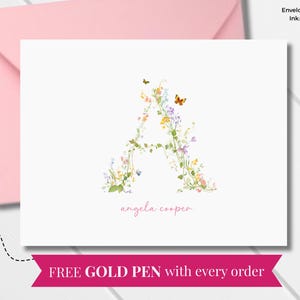 Op de afbeelding: Een gevouwen notitiekaart met een bloemig "A"-ontwerp, een roze envelop en een gouden pen. De notitiekaart bevat de naam "angela cooper" in een sierlijk handschrift. De tekst "FREE GOLD PEN with every order" is ook zichtbaar.