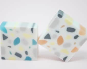 May include: Two square-shaped soap bars with a terrazzo pattern. One bar has blue, gray, and yellow flecks, while the other has orange, gray, and yellow flecks.
