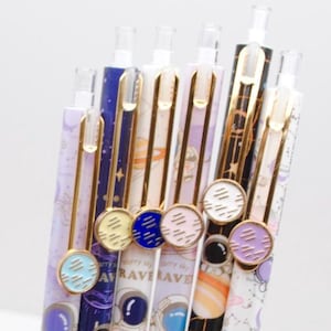 May include: A selection of colourful pens with space-themed designs. The pens feature astronaut illustrations, planets, and the text "RAVE TRAVEL". Each pen has a different colour scheme, including purple, blue, gold, and white, with decorative circular toppers.