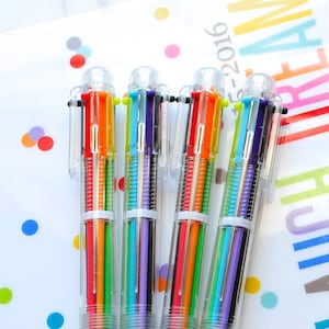 May include: A close-up of five multi-colored pens with clear plastic barrels. The pens are arranged in a row on a white background with colorful polka dots. The text "LIVE, LAUGH, DREAM" is visible on the background.