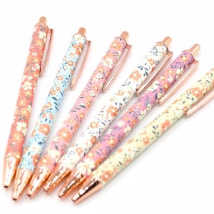 Peut inclure: Six stylos à motifs floraux dans les tons corail, bleu, rose et jaune. Chaque stylo est doté d'un clip et d'une pointe en or rose. Les stylos sont disposés sur une surface blanche, mettant en valeur leurs motifs colorés. Accessoire d'écriture.