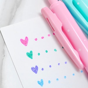 May include: A close-up of colorful pens and a white card with heart and dot designs. The pens are pink and teal, with a pink pen in focus. The card features pink, green, purple, and blue hearts and dots.