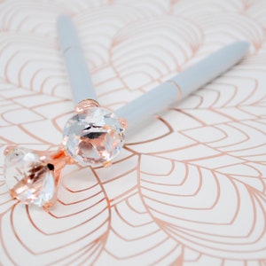 May include: Two light blue pens with clear rhinestone tops. The pens are lying on a white surface with a rose gold pattern.