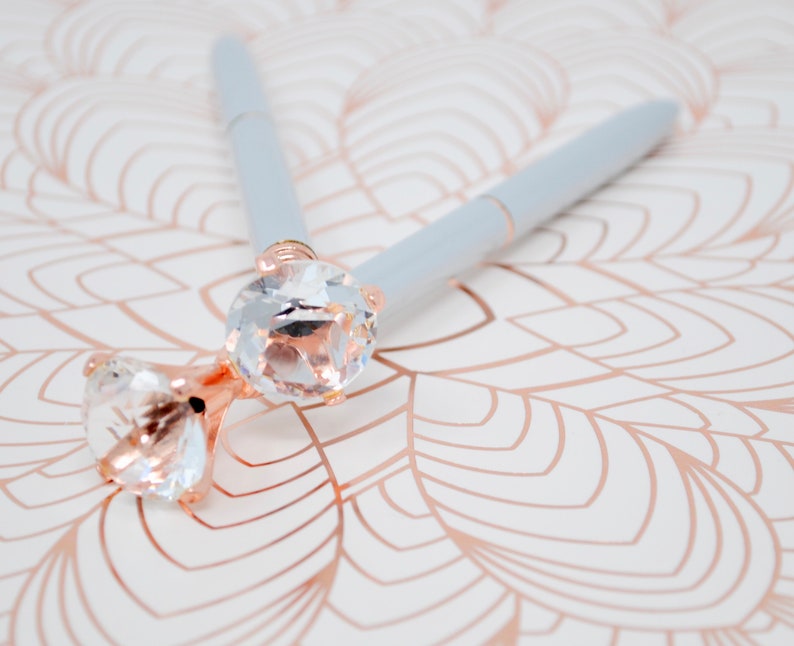 May include: Two light blue pens with clear rhinestone tops. The pens are lying on a white surface with a rose gold pattern.
