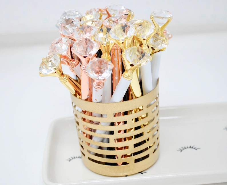 May include: A gold wire basket filled with a variety of pens with clear rhinestone tops. The pens are in gold, rose gold, and white with black polka dots.