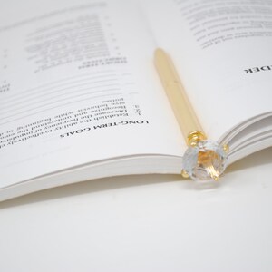 May include: A gold pen with a clear crystal top is resting on an open book. The book is open to a page with the heading "Long-Term Goals" and a list of items.