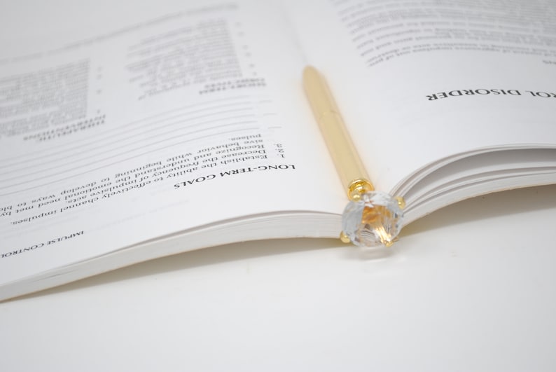 May include: A gold pen with a clear crystal top is resting on an open book. The book is open to a page with the heading "Long-Term Goals" and a list of items.