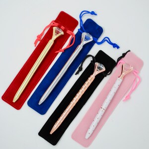 May include: Five diamond-shaped pens in velvet pouches. The pens are gold, silver, rose gold, and white with black polka dots. The pouches are red, blue, black, and pink.