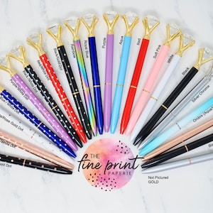 May include: A variety of pens with diamond-shaped tops and different colored barrels. The pens are arranged in a semi-circle on a white marble surface. The text "The Fine Print Paperie" is visible in the center of the image.