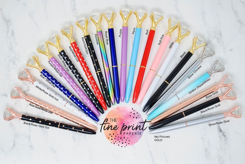 May include: A variety of pens with diamond-shaped tops and different colored barrels. The pens are arranged in a semi-circle on a white marble surface. The text "The Fine Print Paperie" is visible in the center of the image.