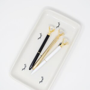 May include: Three pens with gold diamond-shaped tops on a white tray with black eyelash designs. One pen is black, one is white, and one is gold.