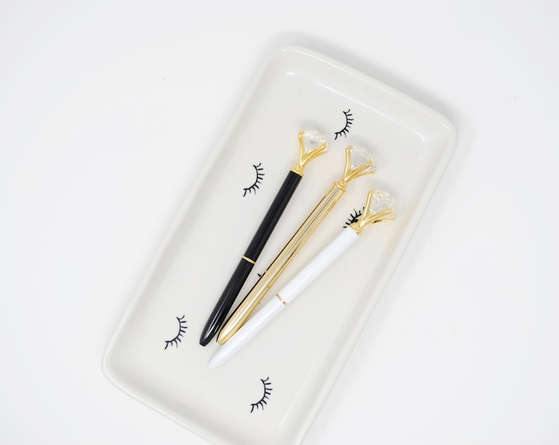 May include: Three pens with gold diamond-shaped tops on a white tray with black eyelash designs. One pen is black, one is white, and one is gold.
