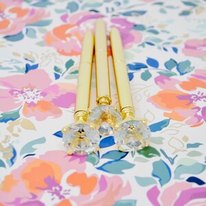 May include: Three gold pens with clear rhinestone tops on a floral background.