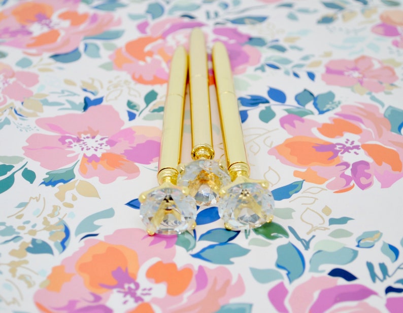 May include: Three gold pens with clear rhinestone tops on a floral background.