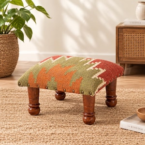 May include: A low footstool featuring a rectangular top upholstered in a woven fabric with a geometric pattern in shades of orange, olive green, and cream. The stool is supported by four dark brown turned wooden legs with brass nailhead trim.