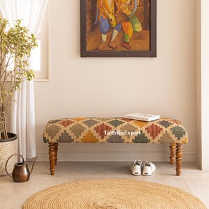 May include: A patterned bench with a rectangular top in green, yellow, red, and brown. The bench has four wooden legs and a curved top. A book and a pair of white shoes are on the bench. A round jute rug is on the floor.