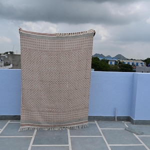 May include: A rectangular textile throw featuring a repeating geometric pattern in dark brown and cream. The fabric has horizontal striped borders at the top and bottom edges, finished with short, frayed fringe detailing along both ends.