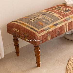May include: A rectangular bench with a patterned textile top in shades of red, blue, and yellow. The bench has four turned wooden legs. The textile features geometric designs. The bench is a decorative furniture piece.