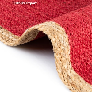 Op de afbeelding: Close-up van een rode jute vloerkleed met een natuurlijke vezelrand. Het vloerkleed heeft een geweven textuur en een gevouwen rand, waardoor de onderkant zichtbaar is. De tekst "TirthikaExport" is zichtbaar in de linkerbovenhoek.