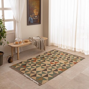 Pode incluir: Um tapete retangular com um padrão geométrico repetitivo de triângulos em tons de verde suave, castanho escuro, bege e laranja queimado. O tapete tem uma textura tecida e franjas curtas nas extremidades mais curtas, com o texto Jaipurrugsexports.